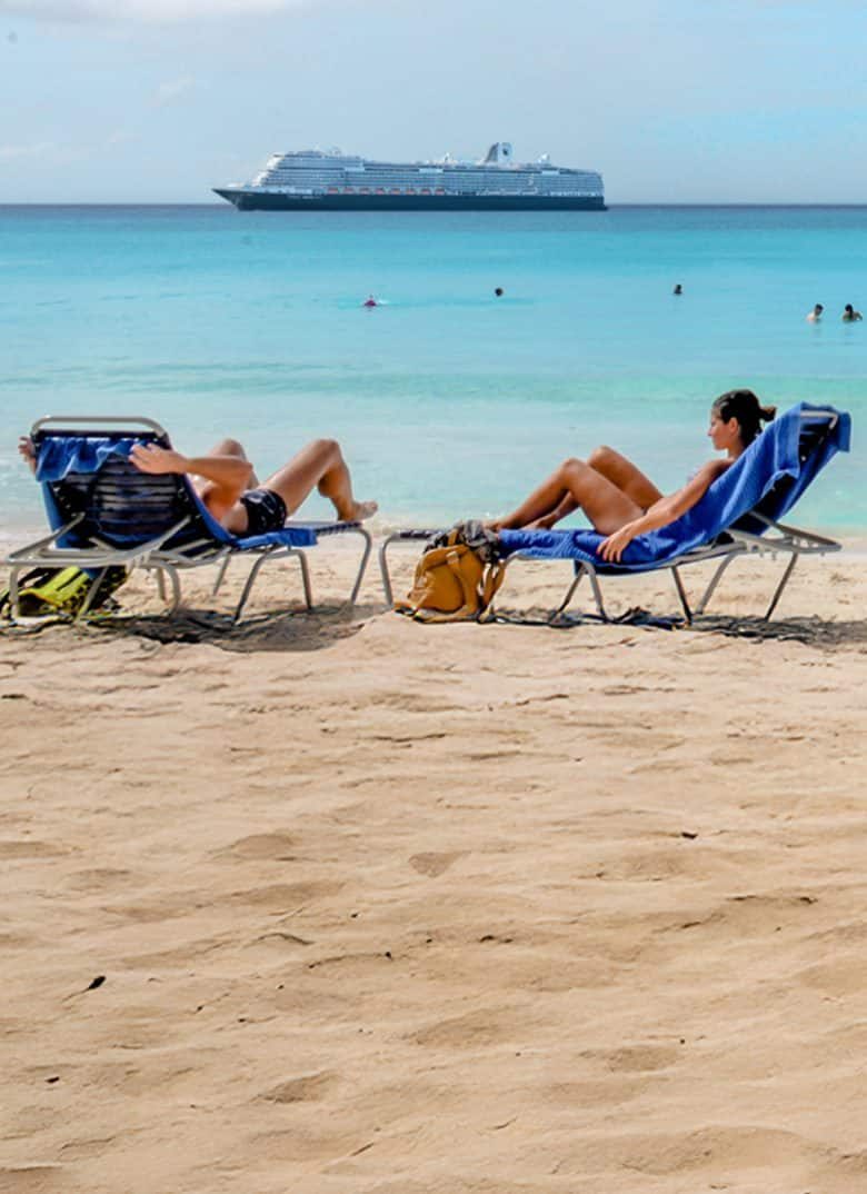 Two guests on blue loungers on the sand at Half Moon Cay with a Holland America ship anchored in the distance